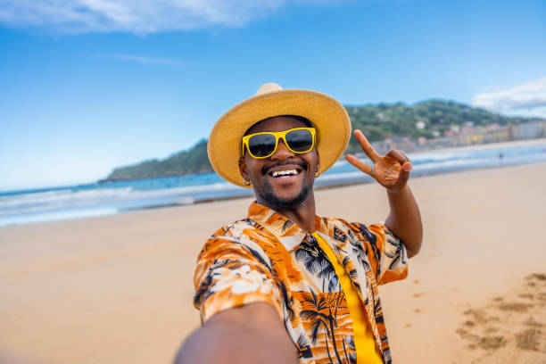 African american man in sun hat, sunglasses and colorful clothes gesturing victory while taking a selfie on beach on vacation.